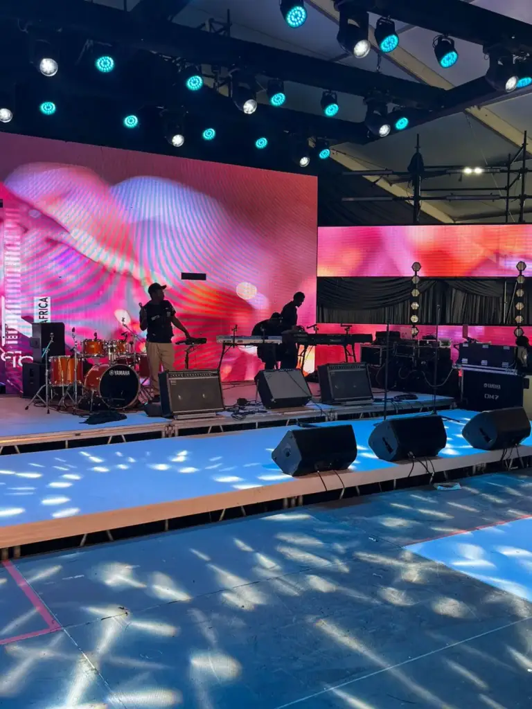 Musicians sound-checking on a wide stage with a Yamaha drum kit, keyboards and wedge monitors under moving-head lights in Kenya