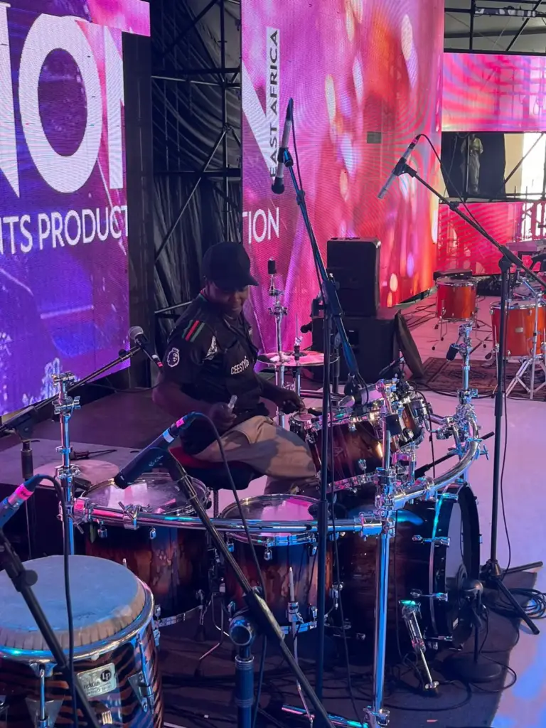 Drummer sound-checking a burst finish drum kit with congas in the foreground on a large tented stage in Kenya