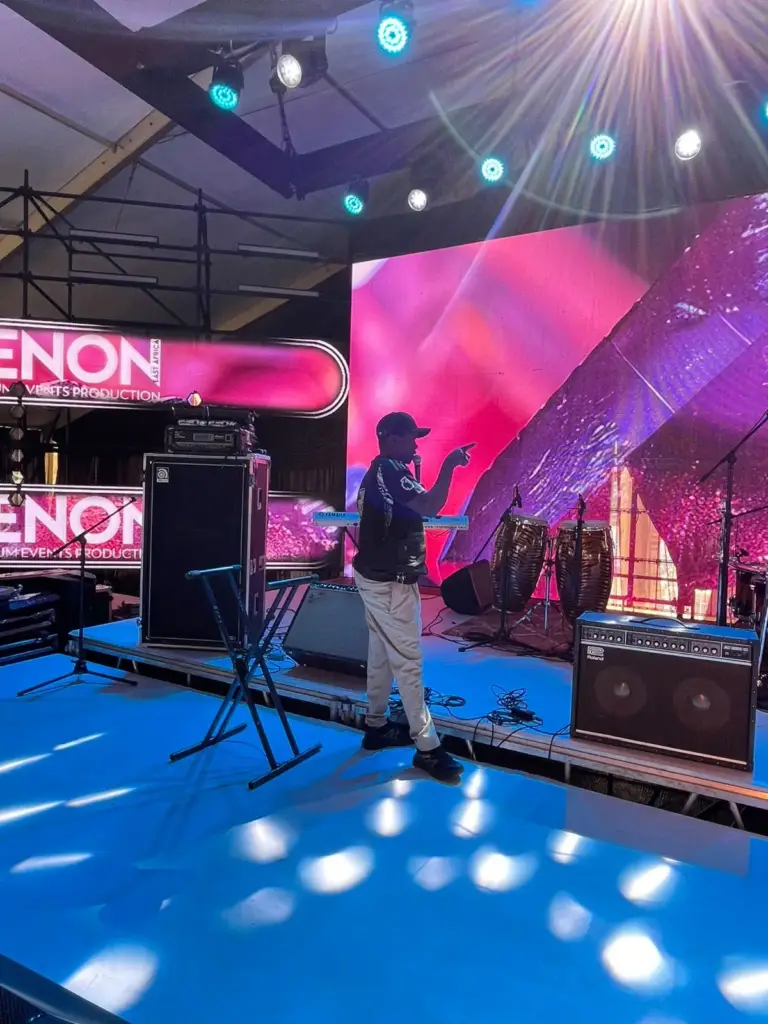Musician directing a sound check beside an Ampeg bass cabinet, congas and a Roland amp on a stage with LED walls in Kenya