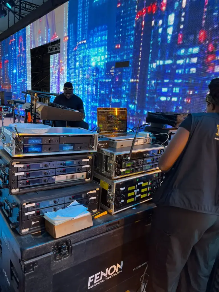 Technician checking racks of wireless microphone receivers at the stage edge in front of a blue LED wall in Kenya