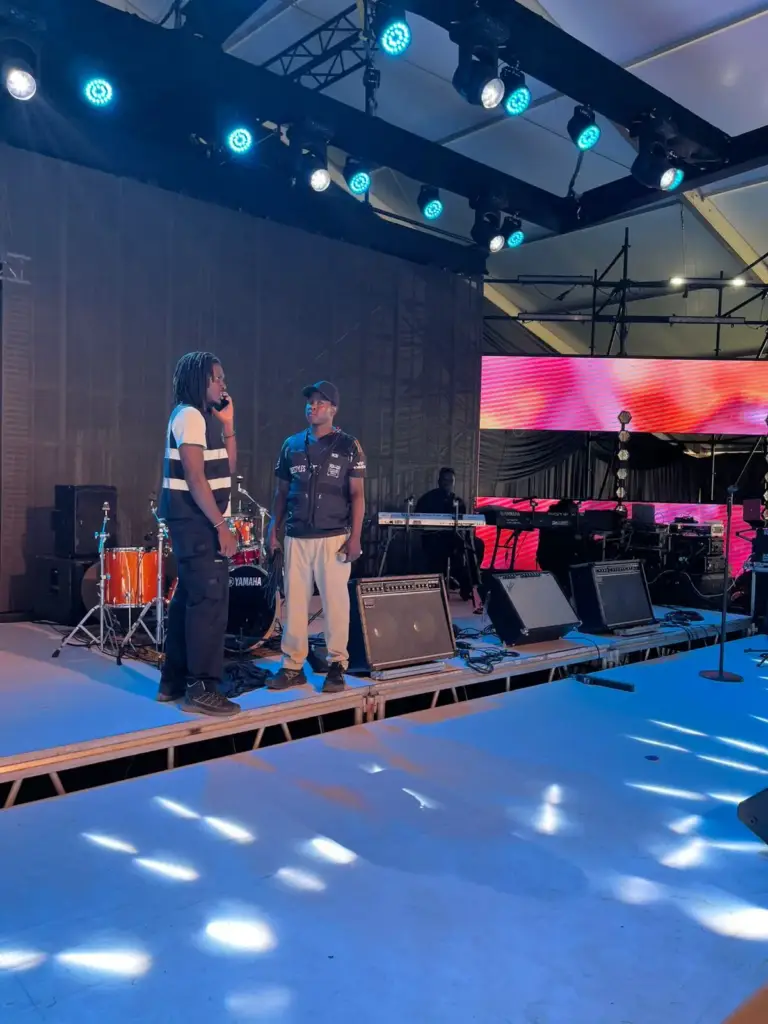 Two crew members on stage beside a Yamaha drum kit and guitar amps while a keyboard player sets up behind them in Kenya