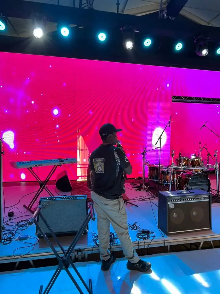 Performer holding a microphone during sound check in front of a pink LED wall, with Roland amps and keyboards on stage in Kenya